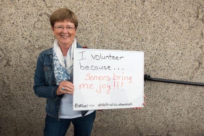 Smiling woman holding up a sign that says "I volunteeer because... Seniors bring me joy!"