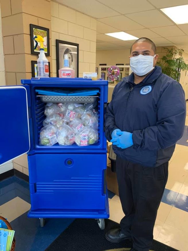 A worker from Neigbhorhood House, wearing a mask and gloves, poses in front of a cabinet full of pre-made meals. 